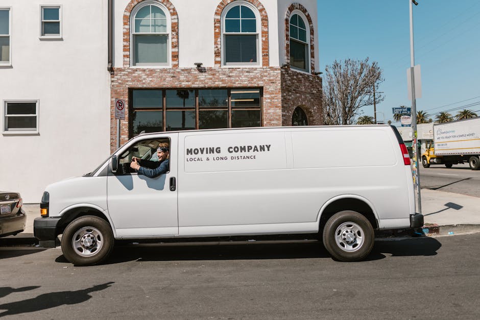 A white moving van with the branding 'MOVING COMPANY LOCAL & LONG DISTANCE' parked on a city street in front of a brick building with large front windows and a second-floor arched window. The van's driver, a man wearing glasses and a dark jacket, is seen smiling and holding the steering wheel from the open driver’s side window. The scene includes additional parked vehicles along the curb, a sidewalk, and a street sign, with a partly cloudy sky overhead. This image captures the loading or transport phase of a home relocation process, with the van ready for furniture transport or packing and moving tasks, characteristic of services provided by companies like Man with Van Surrey specializing in removals.