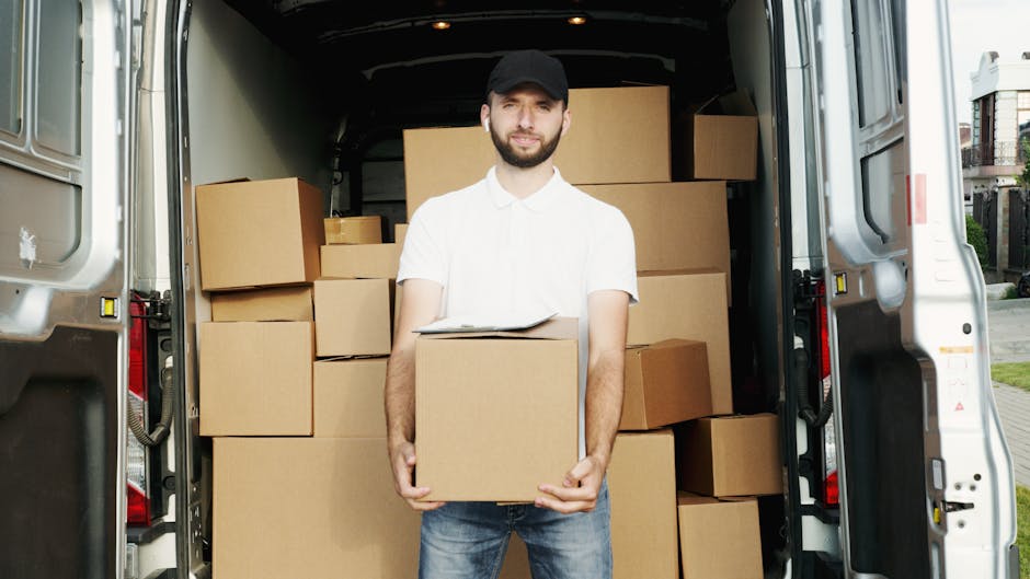 A white moving van with the branding 'MOVING COMPANY LOCAL & LONG DISTANCE' parked on a city street in front of a brick building with large front windows and a second-floor arched window. The van's driver, a man wearing glasses and a dark jacket, is seen smiling and holding the steering wheel from the open driver’s side window. The scene includes additional parked vehicles along the curb, a sidewalk, and a street sign, with a partly cloudy sky overhead. This image captures the loading or transport phase of a home relocation process, with the van ready for furniture transport or packing and moving tasks, characteristic of services provided by companies like Man with Van Surrey specializing in removals.